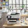 A Canon MAXIFY GX 7050 Multifunktionsdrucker is shown on a wooden desk in a bright, modern office or retail space. The printer is in the foreground, with ink bottles and a calculator on the desk. In the background, there are shelves with items, large windo Produktbild-Vorschau