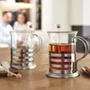 Two glass tea mugs with metal holders and wooden stirrers on a wooden table, with two people blurred in the background. Produktbild-Vorschau