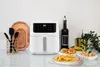 A white air fryer is displayed on a countertop with various food items and kitchen accessories. Voir les images du produit