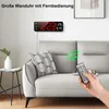 A digital wall clock is mounted on a wall above a sofa, with a hand holding a remote control pointed towards the clock. Produktbild-Vorschau