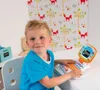 A young boy is sitting at a desk, looking at the camera and interacting with a toy laptop. The background features playful wallpaper with foxes and trees. Náhľad obrázku produktu