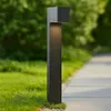 A modern, dark gray outdoor lamp stands next to a paved walkway, with green grass and trees in the background. Produktbild-Vorschau