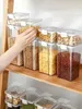 A hand is reaching for a clear plastic food storage container filled with pasta, which is part of a larger set of containers on a wooden shelf. Produktbild-Vorschau