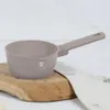 A taupe saucepan with a handle rests on a wooden board next to a folded cloth. Produktbild-Vorschau