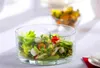 A glass bowl filled with salad, including lettuce, cherry tomatoes, and what appear to be small mozzarella balls, is placed on a white tablecloth. Another blurred bowl of food is visible in the background. Podgląd zdjęcia produktu