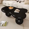A black, cloud-shaped coffee table with a bowl of fruit and a cup of coffee on it, placed on a carpet in front of a white sofa. Voir les images du produit