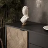 A close-up of a modern sideboard with a decorative bust on top, set against a textured wall and a large plant. Produktbild-Vorschau