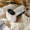 A white lint remover with a digital display sits on a textured beige blanket, next to an open book and some dried grass. Produktbild-Vorschau