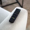 A black remote control rests on the armrest of a light-colored chair. Náhled obrázku produktu