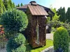 A wooden smoker box with a thermometer attached, situated in a garden setting with manicured bushes and trees. Anteprima dell’immagine prodotto
