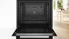 An open black oven with a clean interior, set against a white wall and a glimpse of a dining area in the background. Produktbild-Vorschau