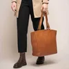 A person wearing a trench coat and black pants, holding a brown leather tote bag. Voir les images du produit