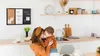 A mother and child are embracing in a kitchen setting, with a bulletin board on the wall and kitchen appliances in the background. Podgląd zdjęcia produktu