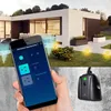 A hand holding a smartphone displaying a smart home app, with a modern house and garden in the background, and a smart plug in the foreground. Náhled obrázku produktu