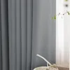 A close-up view of a gray fabric curtain with vertical pleats, partially covering a window with sheer white curtains behind it. A small green plant branch is visible to the right, and a gift box with a ribbon is in the foreground. Produktbild-Vorschau