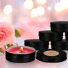 A lit rose-scented candle in a black tin is in the foreground, with several other black tin candles stacked behind it. A pink rose is to the left of the candle. The background is blurred with bokeh lights. Produktbild-Vorschau