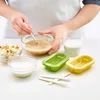 Hands mixing ingredients in a bowl, with ice pop molds and ingredients on a white surface. Produktbild-Vorschau