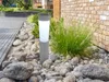 A solar-powered garden light is placed in a rock garden next to a brick wall and a paved walkway. Produktbild-Vorschau