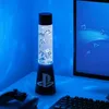 A PlayStation-themed lava lamp sits on a desk next to a computer monitor and keyboard, with a game controller in the foreground. Podgląd zdjęcia produktu