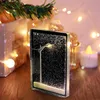 A decorative snow globe with a street lamp illuminated, set against a festive, blurred background with Christmas lights and greenery. Produktbild-Vorschau
