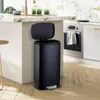 A black pedal trash can is shown in a modern kitchen setting, next to a kitchen island and oven. Produktbild-Vorschau