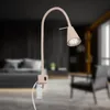 A beige LED wall lamp with a flexible gooseneck is mounted on a wall in a bedroom setting. The lamp is positioned next to a bed with pillows and a dark headboard, and a chair is visible in the background. Náhled obrázku produktu