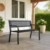 A black metal garden bench is placed on a wooden deck in front of a white house with windows. A large potted plant with white flowers is to the right of the bench, and a green hedge is in the background. Produktbild-Vorschau