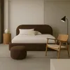 A modern bedroom with a brown upholstered bed, a wooden chair, a side table, and a floor lamp. Produktbild-Vorschau
