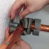 Hands are shown installing pipe insulation onto a copper pipe. The insulation is gray and segmented, and clips are visible. The pipe has text printed on it. Produktbild-Vorschau