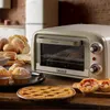 A vintage-style toaster oven is shown on a wooden table, surrounded by freshly baked goods like bread, muffins, and a pie. Náhled obrázku produktu