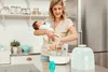 A woman is holding a baby and using a bottle sterilizer in a kitchen. Podgląd zdjęcia produktu