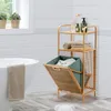 A bathroom scene featuring a wooden shelf unit with a fold-out laundry basket and towels, next to a bathtub. Produktbild-Vorschau