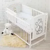 A white baby crib with bedding featuring a teddy bear and golden stars pattern, next to a white teddy bear toy and a fluffy rug. Anteprima dell’immagine prodotto