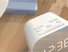 A close-up of a white digital alarm clock with buttons on top, placed on a wooden surface next to some books. Podgląd zdjęcia produktu