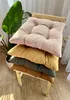 A stack of four colorful seat cushions on a wooden chair, with a large green plant in the background. Produktbild-Vorschau