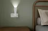 A bedside scene with a wall-mounted night light and outlet, and a bed with a pillow and headboard. Produktbild-Vorschau