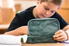 A young boy is sitting at a desk, looking down at an open pencil case. Produktbild-Vorschau