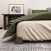 A close-up view of a bed with a green blanket and a beige comforter, showing a black bed riser at the bottom corner. Produktbild-Vorschau