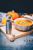 A nutmeg grinder and a bowl of pumpkin soup are arranged on a wooden board, with pumpkins in the background. Produktbild-Vorschau