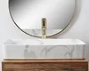 A rectangular bathroom sink with a marble pattern, a gold faucet, and a round mirror in the background, placed on a wooden cabinet. Anteprima dell’immagine prodotto
