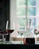 A collection of RIEDEL wine glasses and a decanter filled with red wine, set on a dark countertop with a blurred background of a window and greenery. Produktbild-Vorschau