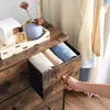 A hand is opening a drawer of a dresser with clothes inside. There are also decorative items on top of the dresser. Náhled obrázku produktu