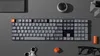 A top-down view of a mechanical keyboard and other desk accessories arranged on a gray surface. Produktbild-Vorschau