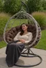 A woman is relaxing in a hanging egg chair outdoors, covered with a blanket. Voir les images du produit
