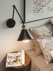 A bedside table with a magazine next to a bed with pillows, illuminated by a wall-mounted lamp. Náhled obrázku produktu