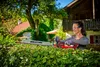 A woman is using a hedge trimmer to trim a hedge in a garden, with a house and trees in the background. Produktbild-Vorschau