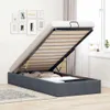 A bed frame with the storage compartment open, revealing the slatted base and some bedding inside. The bed is in a room with a window and wall art in the background. Produktbild-Vorschau