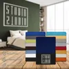 A stack of colorful fitted sheets is displayed in front of a bedroom scene with a bed and a wall with a logo. Produktbild-Vorschau