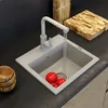 A kitchen sink with a faucet running water into it, and two red tomatoes inside. A wooden cutting board and bowls are on the counter. Produktbild-Vorschau