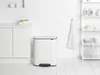 A white Brabantia Bo Mülleimer (trash can) with a black pedal is placed on a white wooden floor next to a white cabinet in a clean, minimalist kitchen setting. A white towel hangs on the wall behind it, and a white shelf unit with various kitchen items is  Náhľad obrázku produktu